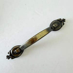 May include: An antique-style drawer pull with a rectangular bar and decorative circular ends. The metal hardware has a tarnished, aged appearance with a mix of silver and gold tones. The ends feature ornate, scroll-like detailing.