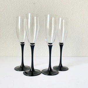 May include: Four champagne flutes with clear glass bowls and black stems and bases. The flutes are arranged in a group, showcasing their elegant design. The black bases provide a striking contrast to the clear glass.