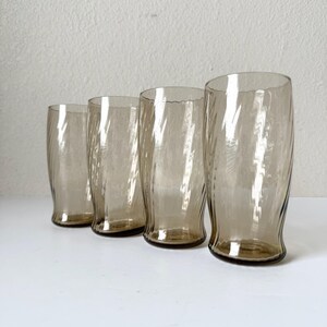 May include: A set of four tall drinking glasses made of translucent smoke-colored glass. Each glass features a distinct vertical ribbed or fluted texture that creates a wavy, undulating pattern along the exterior surface of the cylindrical body.