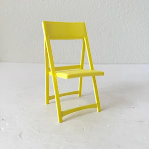 以下が含まれることがあります： 鮮やかな黄色の小さな折りたたみ椅子。 椅子は長方形の背もたれと座面があり、角度の付いた脚とシンプルなフレームが付いています。 椅子はしっかりとした素材でできており、収納のために平らに折りたたむことができます。 背景はプレーンな白い表面です。
