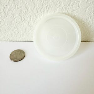 May include: A circular, translucent white plastic lid with a smooth, flat surface and a raised outer rim. The lid is positioned next to a silver-colored United States quarter coin, which serves as a visual reference for the scale of the object.