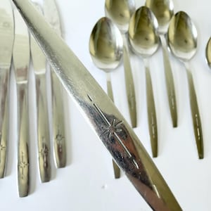 May include: A collection of stainless steel flatware including several spoons and knives. Each piece features a polished silver finish and a distinct engraved starburst pattern on the handle. The items are arranged in a group with one spoon handle prominently displayed in the foreground showing the etched starburst detail.