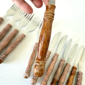 K&ouml;nnte beinhalten: Ein Set aus Edelstahlmessern und -gabeln mit Holzgriffen. Die Griffe haben eine hellbraune, lackierte Oberfl&auml;che. Das Besteck ist in klarem Kunststoff verpackt. Die Aufnahme erfolgt aus leicht erh&ouml;hter Perspektive.