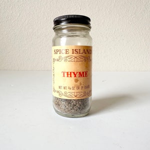 Może przedstawiać: Szklany słoik z tymiankiem z czarną pokrywką. Etykieta głosi "SPICE ISLAND" i "THYME" w czerwonych literach. Waga netto wynosi 21 gramów. Słoik jest wypełniony suszonym tymiankiem.