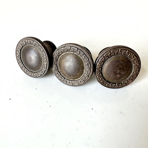 May include: Three antique metal cabinet knobs. Each knob has a circular design with a raised, patterned border around a central, slightly domed disc. The knobs have a tarnished, aged appearance, suggesting they are vintage or reclaimed.