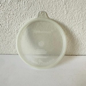 Op de afbeelding: Een helder, rond Tupperware-deksel met een klein handvat. Het deksel is gemaakt van doorschijnend plastic en heeft het Tupperware-logo en andere tekst. Het deksel is ontworpen om op een container te passen en heeft een iets verhoogde rand.