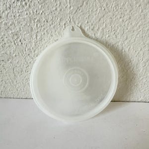 May include: A clear, round Tupperware lid with a small handle. The lid has the Tupperware logo and other text printed on it. The lid is made of a translucent plastic material.
