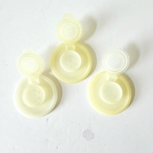 May include: Three pale yellow plastic bottle caps with attached hinged lids. Each cap has a circular base with a smaller, perforated circle in the center. The lids are open, revealing the interior.