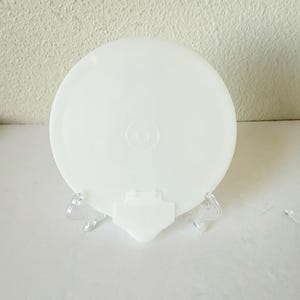 May include: A white, round Tupperware lid, displayed on a clear plastic stand. The lid has a slightly domed shape with a circular design in the center. The stand has a diamond-shaped base and two curved supports.