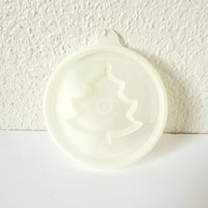 Puede incluir: Molde redondo de plástico blanquecino para adornos con diseño de árbol de Navidad. El molde tiene un pequeño lazo en la parte superior para colgar. El diseño es una forma de árbol simple en relieve.