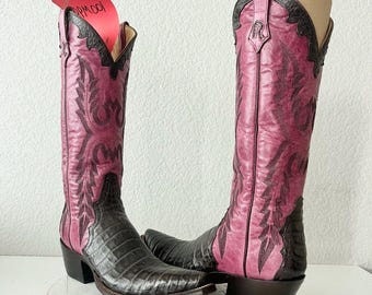 May include: A pair of tall cowboy boots featuring a magenta shaft with black decorative stitching and a black textured foot. A red tag with the handwritten text RPM001 is attached to the top of the left boot. The boots have a pointed toe.