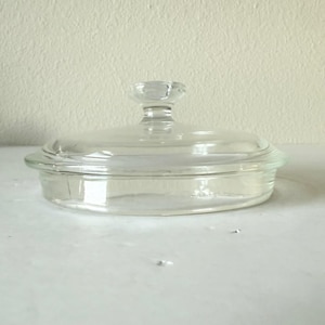 May include: A clear glass casserole dish with a matching lid. The lid has a small, rounded handle. The dish and lid are made of transparent glass, allowing visibility of the contents. The dish is round with a slight lip.