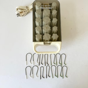 May include: A vintage General Electric MiniSetter hair curler set. The set includes a cream-colored case with a clear lid, a handle, and a collection of gray curlers. Metal clips in black and green are also included.