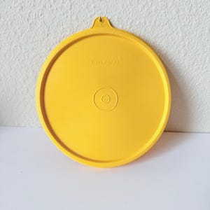 May include: Bright yellow Tupperware lid with a circular design. The lid has a small loop at the top and the brand name is printed on the surface. The lid has a raised edge and a circular indentation in the center.