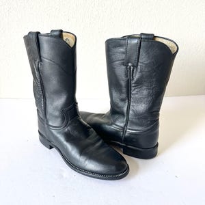 Könnte beinhalten: Ein Paar wadenhohe Stiefel aus schwarzem Leder mit abgerundeter Zehenpartie, niedrigem Blockabsatz und vertikalen Zuglaschen am Schaft. Die Stiefel haben sichtbare Nähte entlang der Sohle und eine glatte, matte Oberfläche des Außenleders.