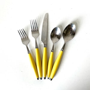 May include: A set of five stainless steel utensils: two forks, a knife, and two spoons. Each piece has a bright yellow handle with a silver band at the base. The utensils are arranged on a white surface.