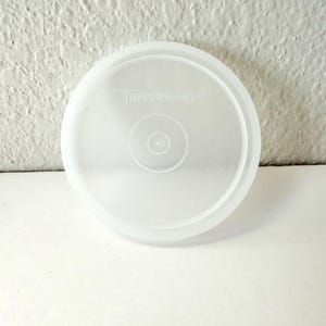 May include: A clear, round Tupperware lid. The lid is made of translucent plastic and features the Tupperware logo. The lid has a slightly raised center with concentric circles. The lid is designed to fit a container.