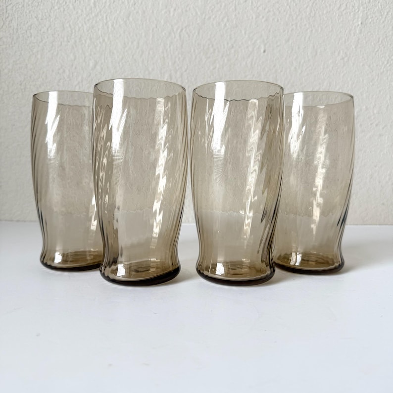 May include: A set of four tall drinking glasses made of translucent smoke-colored glass. Each glass features a distinct vertical swirl pattern that creates a rippled texture along the exterior surface, with a slightly flared rim and a weighted base.