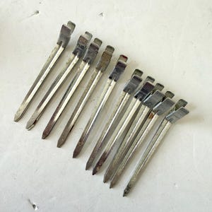 May include: A collection of vintage silver metal hair clips, arranged diagonally on a white surface. The clips have a rectangular top and a pointed end. Some clips show signs of age and wear.