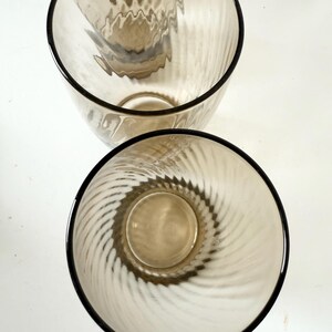 May include: Two drinking glasses made of translucent, light brown tinted glass. Each glass features a distinct vertical ribbed or fluted texture that spirals slightly along the interior walls, creating a repeating pattern of light and shadow.