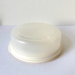 May include: A round, semi-translucent off-white plastic cake carrier with a flat top and a matching circular base. The container features a smooth, minimalist design with a secure locking rim, providing a clear view of the interior space.