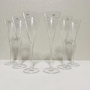 May include: A set of six clear champagne flutes. Each glass has a tall, slender stem and a wide, conical bowl. The glasses are arranged in a row, with the bases resting on a white surface. The brand name is visible on each glass.