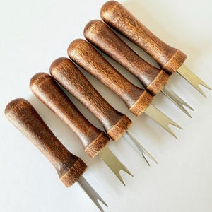 May include: A set of five corn cob holders, each featuring a cylindrical, dark brown wooden handle with a visible wood grain texture and two sharp, silver-toned metal prongs extending from the base for securing food items during consumption.