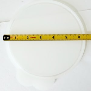 May include: A round, translucent white plastic lid with a small tab at the bottom edge. A yellow Stanley measuring tape is placed across the center, showing the lid has a diameter of approximately 6.5 inches from edge to edge.