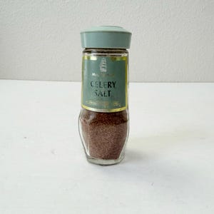 Op de afbeelding: Een vintage glazen selderijzoutstrooier met een lichtgroene deksel en een groen etiket. Het etiket bevat de witte woorden "CELERY SALT". De strooier is gevuld met een bruin, korrelig kruid.