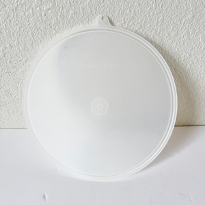 May include: A clear, round plastic lid with a small handle. The lid has a slightly textured surface and a raised rim. The center of the lid features a circular design. The lid is designed to fit a container.