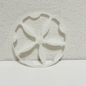 May include: A white plastic circular object with a floral design. The object has six petal-shaped elements radiating from a central point, surrounded by a scalloped edge. The object is set against a white background.