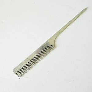 May include: A silver-colored metal comb with a long, pointed handle. The comb has fine teeth along one edge and is designed for hair styling. The comb is shown against a white background.