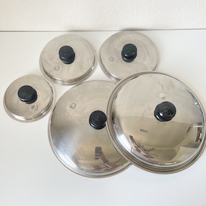 May include: Five stainless steel pot lids of varying sizes are arranged on a white surface. Each lid has a black knob in the center. The lids reflect the surrounding environment.