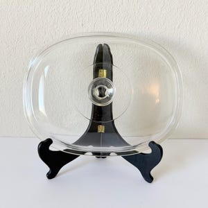 May include: A clear, oval-shaped glass lid with a central knob, displayed on a black decorative stand. The lid is transparent, allowing a view of what it would cover. The stand has a curved design and is made of black wood.