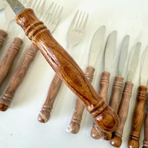 K&ouml;nnte beinhalten: Ein Set aus Edelstahlmessern und -gabeln mit Holzgriffen. Die Griffe haben eine satte, braune Farbe und eine gl&auml;nzende Oberfl&auml;che. Das Besteck ist in einer ordentlichen Reihe angeordnet, was auf ein Essgeschirr-Set hindeutet.