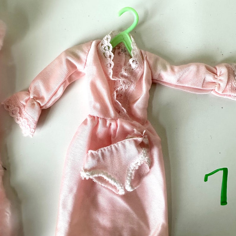 Op de afbeelding: Een roze satijnen nachtjapon met kant en bijpassende roze satijnen onderbroek. De nachtjapon hangt aan een groene plastic hanger.