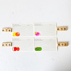 May include: Four recipe cards with lined paper and fruit illustrations, including a lemon, grapes, apples, and watermelon. Wooden letter tiles spell out "A 14", "A 15", "A 16", and "A 17". The cards have the text "Here's what's cookin'" and "Recipe from the kitchen of".