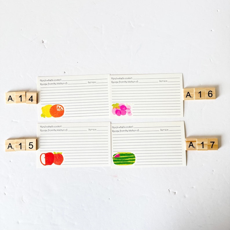 May include: Four recipe cards with lined paper and fruit illustrations, including a lemon, grapes, apples, and watermelon. Wooden letter tiles spell out "A 14", "A 15", "A 16", and "A 17". The cards have the text "Here's what's cookin'" and "Recipe from the kitchen of".