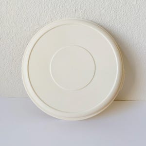 Könnte beinhalten: Ein runder, cremefarbener Kunststoffdeckel mit erhöhtem Rand und einer zentralen, kreisförmigen Vertiefung. Der Deckel ist für einen Behälter bestimmt und hat ein schlichtes, funktionales Design.