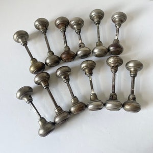 May include: A collection of antique door knobs with a tarnished, metallic finish. The knobs have a rounded shape and are attached to long, slender stems. The metal shows signs of age and wear, with variations in color and texture.