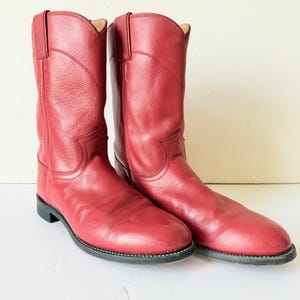 Könnte beinhalten: Ein Paar wadenhohe Cowboystiefel aus glattem rotem Leder. Die Stiefel haben ein klassisches Schlupfdesign mit seitlichen Zuglaschen, eine abgerundete Zehenpartie und einen niedrigen Absatz. Das Leder weist eine feine Narbung und sichtbare Nähte auf.