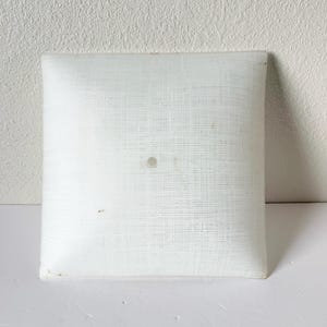 May include: Square, white light fixture with a textured, woven-like surface. The fixture has a single, small, circular detail in the center. The edges are slightly rounded. The background is a white wall and surface.
