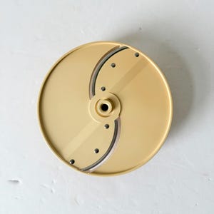 May include: A beige food processor slicing blade. The circular blade has two curved cutting edges and several small holes. The center has a circular hub. The blade is designed for slicing vegetables and other foods.