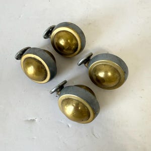 May include: Four antique casters with a brass-colored, rounded center and a gray metal rim. Each caster has a small, silver-colored mounting plate. The casters are arranged in a circular pattern.