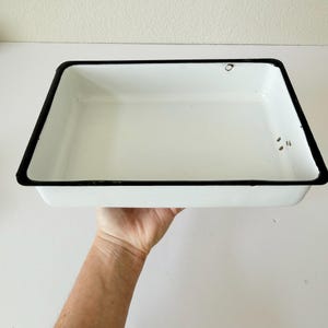 May include: A rectangular white enamel tray with a black border. The tray is held up by a person's hand. The tray has a few small imperfections. This vintage tray is suitable for kitchen or home decor.