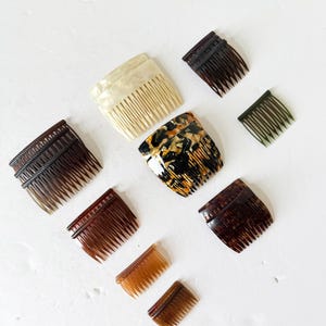 May include: A collection of nine hair combs in various sizes and colors, including cream, dark brown, tortoiseshell, and olive green. The combs feature multiple teeth and are arranged in a scattered pattern, showcasing different textures and finishes.