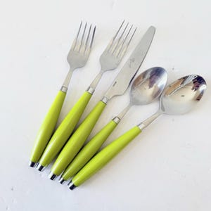 May include: A set of stainless steel flatware with bright green handles. The set includes two forks, a knife, and two spoons. The handles are a solid, glossy green, and the metal parts are silver. The flatware is arranged on a white surface.