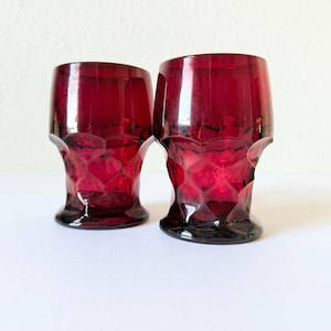 May include: Two ruby red drinking glasses with a textured, geometric pattern. The glasses have a short, rounded shape with a flared base and a thick rim. The glasses are made of a transparent material, allowing light to pass through.