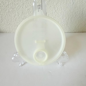 May include: A circular, semi-translucent off-white plastic lid featuring a raised rim and a central circular opening with a small tab. The lid is designed with a smooth, flat surface and a slightly thicker outer edge for a secure fit.