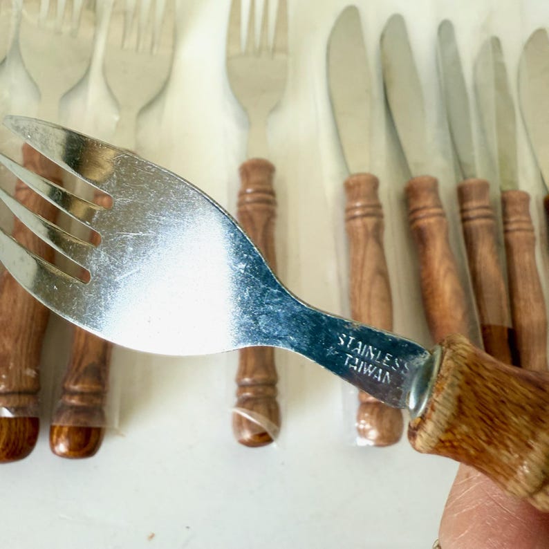 K&ouml;nnte beinhalten: Ein Set aus Edelstahlgabeln und -messern mit Holzgriffen. Die Gabel im Vordergrund hat einen gl&auml;nzenden, silbernen Kopf und einen braunen Holzgriff. Der Text "STAINLESS TAIWAN" ist sichtbar.